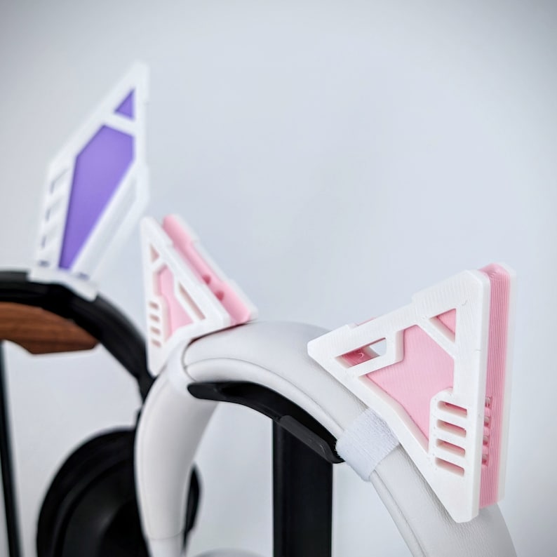 Pode incluir: Fones de ouvido brancos com acess&oacute;rios de orelhas de gato intercambi&aacute;veis em rosa, roxo e branco. As orelhas de gato t&ecirc;m um design geom&eacute;trico e s&atilde;o presas aos fones de ouvido com uma tira de velcro. Os fones de ouvido est&atilde;o em um suporte preto.