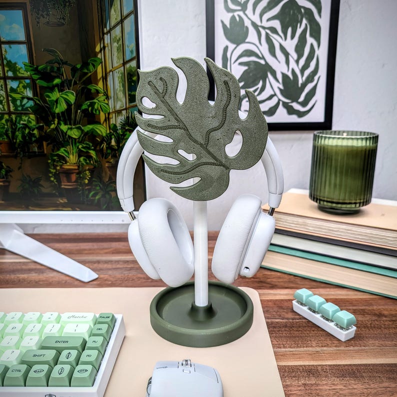 May include: A green monstera leaf-shaped headphone stand with a white base and two white headphones hanging from it. The stand is on a light brown wooden surface with a white keyboard and a white mouse.