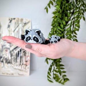 May include: Two gray and black raccoon figurines, one larger than the other, are lying on a hand. The larger raccoon is on the left and the smaller raccoon is on the right.