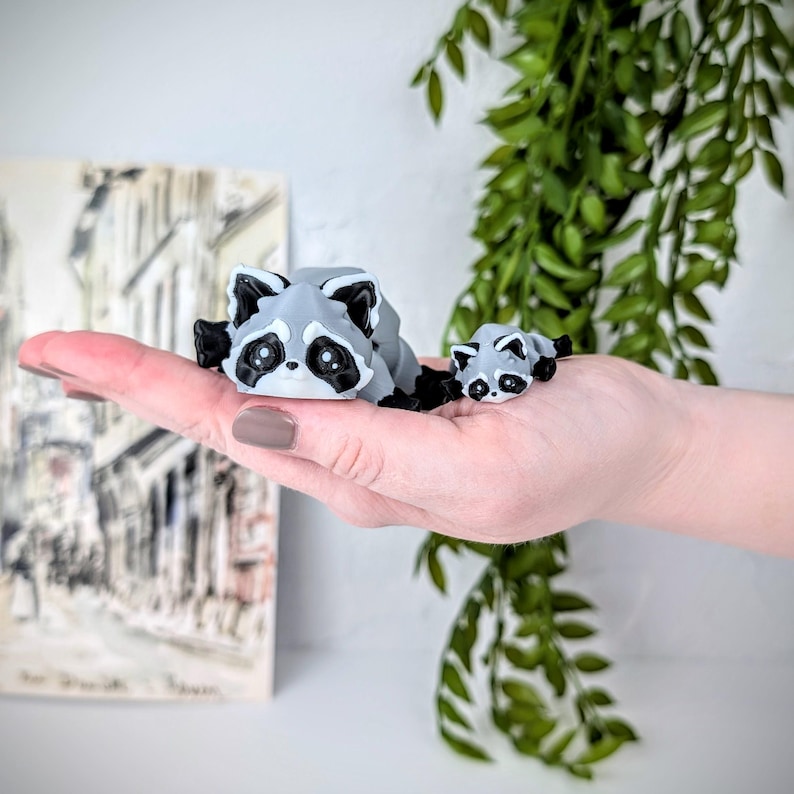May include: Two gray and black raccoon figurines, one larger than the other, are lying on a hand. The larger raccoon is on the left and the smaller raccoon is on the right.