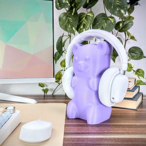 May include: A purple gummy bear shaped headphone stand. The stand is made of plastic and has a white pair of headphones resting on it.