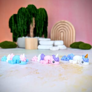 May include: Three small unicorn figurines, each with a different color scheme. The unicorn on the left is blue and white, the middle unicorn is pink and purple, and the unicorn on the right is white and blue. The unicorns are all lying down with their heads turned to the side.