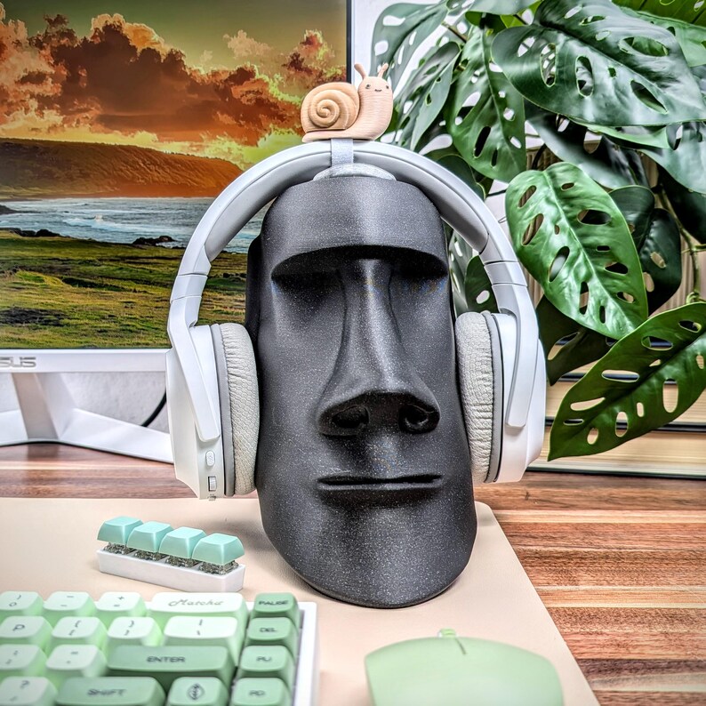 May include: White wireless headphones with a black moai statue on top. The headphones are on a wooden surface with a green plant in the background.