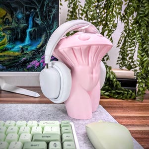 May include: A pink ceramic sculpture shaped like a mushroom with a white pair of headphones on top. The sculpture is on a desk with a green keyboard and a green mouse.