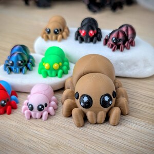 May include: A collection of colorful, 3D-printed spider figurines in various colors, including brown, black, green, blue, pink, and red. Each spider has large, expressive eyes and detailed body features. The spiders are displayed on a white stone and a wooden surface.