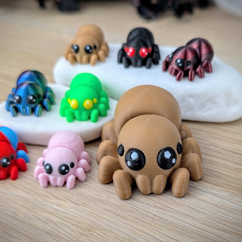 May include: A collection of colorful, 3D-printed spider figurines in various colors, including brown, black, green, blue, pink, and red. Each spider has large, expressive eyes and detailed body features. The spiders are displayed on a white stone and a wooden surface.