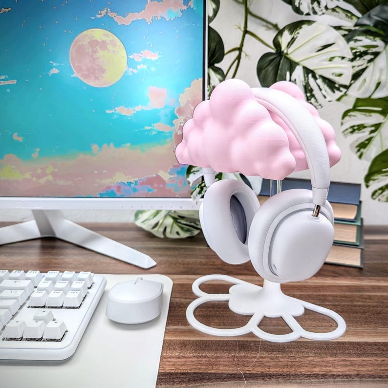K&ouml;nnte beinhalten: Ein wei&szlig;er Kopfh&ouml;rerst&auml;nder mit einem rosafarbenen Wolken-Design. Der St&auml;nder hat eine blumenf&ouml;rmige Basis und ist f&uuml;r das Aufstellen von Kopfh&ouml;rern konzipiert.