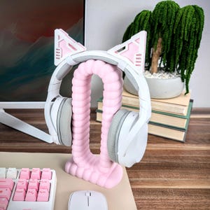 Pode incluir: Um fone de ouvido branco e cinza com orelhas de gato rosa repousa sobre um suporte de fone de ouvido rosa. O suporte tem um design curvo e canelado. Um teclado branco com teclas rosa e um mouse branco est&atilde;o em primeiro plano.