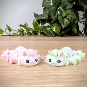 May include: Two small white ceramic figurines of cows with pink and green accents. The cows are lying down with their heads turned to the side.
