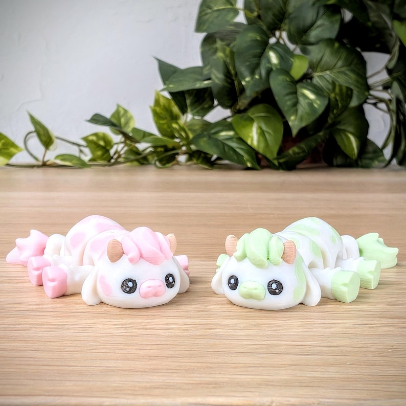 May include: Two small white ceramic figurines of cows with pink and green accents. The cows are lying down with their heads turned to the side.