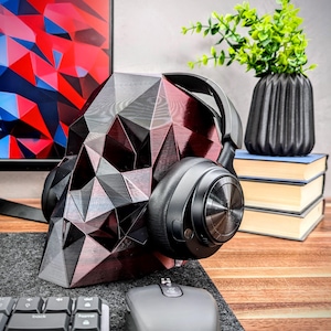 May include: A black and red geometric skull-shaped headphone stand with a pair of black over-ear headphones resting on it. The stand is on a wooden surface with a computer mouse and keyboard in the background.
