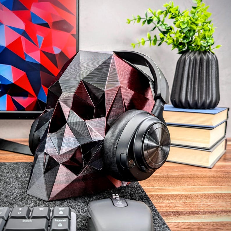 May include: A black and red geometric skull-shaped headphone stand with a pair of black over-ear headphones resting on it. The stand is on a wooden surface with a computer mouse and keyboard in the background.