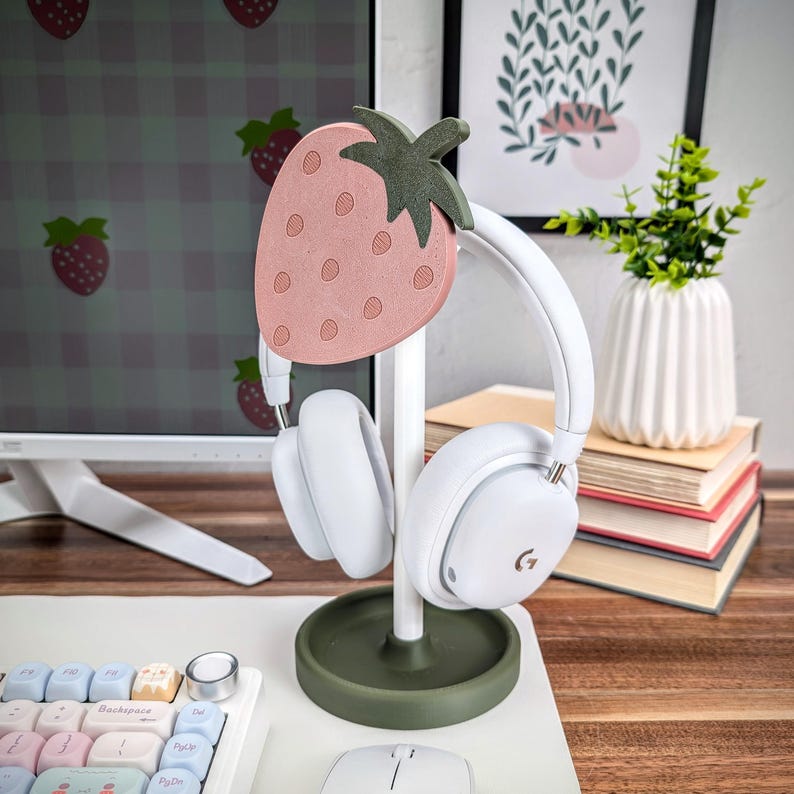 May include: A white headphone stand with a pink and green strawberry design. The stand is made of wood and has a round base. The headphones are white with a black logo on the earcups.