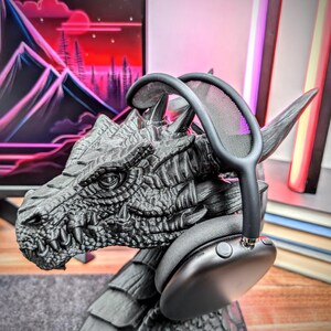 May include: Black headphones with a mesh headband resting on a dragon-shaped stand. The stand is made of a dark gray material and has a detailed dragon head.