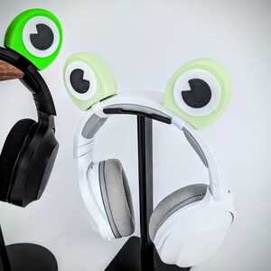 May include: A white headset with green frog-shaped earcups on a black stand. The headset has a white headband and large, round earcups.