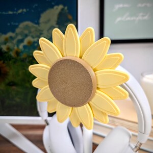 K&ouml;nnte beinhalten: Ein gelber, sonnenblumenf&ouml;rmiger Kopfh&ouml;rerst&auml;nder mit einem strukturierten braunen Zentrum. Der St&auml;nder ist aus Kunststoff und wurde entwickelt, um Kopfh&ouml;rer zu halten.