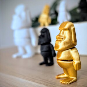 May include: A gold-colored figurine of a person with a large head and a simple body. The figurine is standing on a wooden surface.