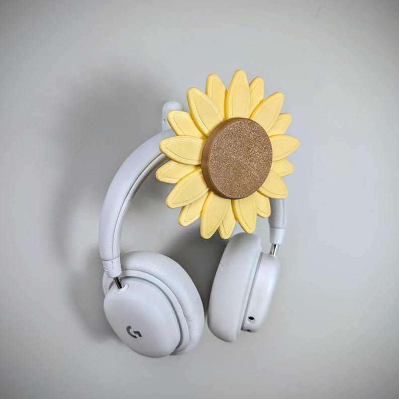 K&ouml;nnte beinhalten: Wei&szlig;e Kopfh&ouml;rer mit einem Kopfh&ouml;rerhalter in Form einer Sonnenblume. Die Sonnenblume ist gelb mit einer braunen Mitte.