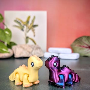 Op de afbeelding: Twee kleine, beweeglijke dinosaurusbeeldjes. E&eacute;n is geel met perzikkleurige accenten, de andere is paars met blauwe en zwarte accenten. Beide hebben grote, zwart-witte ogen en een rij ruggengraat. De achtergrond is een verloop van roze en oranje.