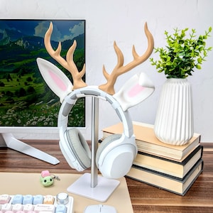 May include: White headphone stand with a deer antler and bunny ear design. The stand is made of metal and has a white base. The headphones are white with gray earcups.
