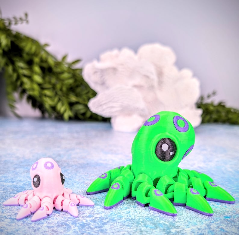 May include: Two octopus figurines, one green and one pink, with purple accents. The green octopus is larger and has a more detailed design. Both octopuses have large, round eyes.
