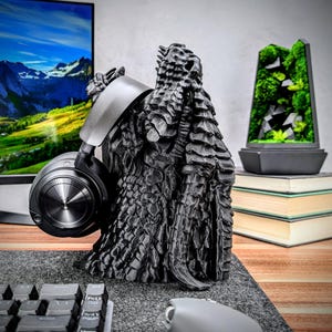 May include: A black 3D printed dragon head headphone stand. The stand is designed to hold a pair of headphones with the earcups resting on the dragon's wings. The stand is on a desk with a keyboard and mouse.