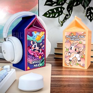 May include: Two colorful milk carton shaped headphone stands. The stand on the left is purple and blue with a cartoon cow and the text "Space Milk". The stand on the right is orange with a cartoon cow and the text "Peaches and Cream".