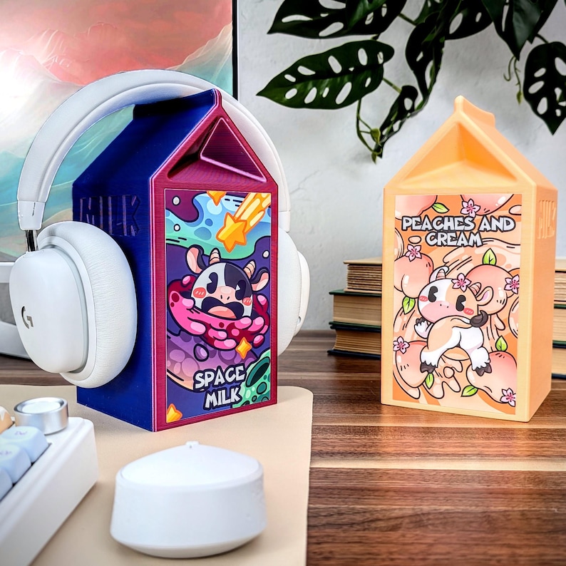 May include: Two colorful milk carton shaped headphone stands. The stand on the left is purple and blue with a cartoon cow and the text "Space Milk". The stand on the right is orange with a cartoon cow and the text "Peaches and Cream".