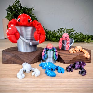 May include: A set of six colourful crab figurines, each holding a small bucket. The crabs are made of plastic and have a variety of colours, including red, pink, blue, orange, and purple.