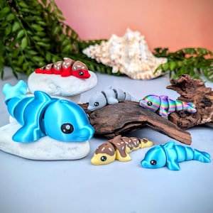 May include: A collection of colorful, 3D-printed dolphin figurines. The dolphins are in various colors, including blue, gray, red, and rainbow. Some are resting on white stones and a piece of driftwood. The figurines have cartoon-like features.