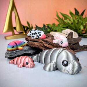 May include: A selection of small manatee figurines in various colours. The figurines include rainbow, pink, and silver. Some have patterns, such as black spots. The manatees are displayed on a piece of driftwood and a dark grey stone.