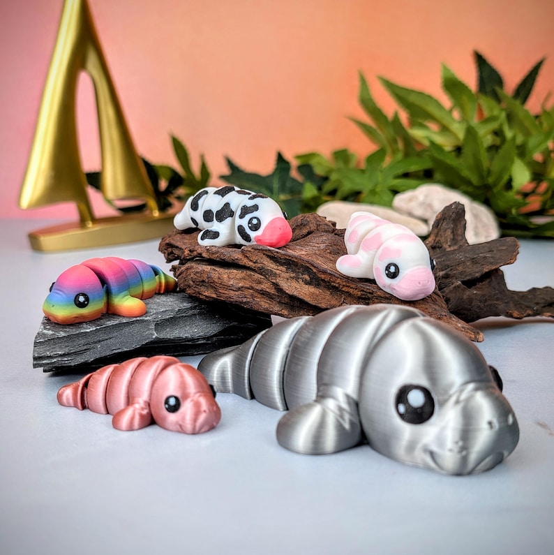 May include: A selection of small manatee figurines in various colours. The figurines include rainbow, pink, and silver. Some have patterns, such as black spots. The manatees are displayed on a piece of driftwood and a dark grey stone.