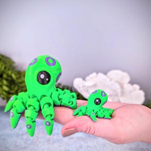 May include: Two green octopus figurines with purple accents. The larger octopus has eight tentacles and the smaller octopus has six tentacles.