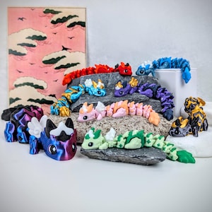 K&ouml;nnte beinhalten: Eine Sammlung farbenfroher, beweglicher Drachenfiguren in verschiedenen Farben, darunter Rot, Blau, Lila und Gr&uuml;n. Einige Drachen haben Fl&uuml;gel und sind auf Felsen drapiert. Im Hintergrund befindet sich ein rosa-roter Druck einer Landschaft mit Wolken.