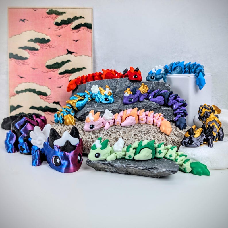K&ouml;nnte beinhalten: Eine Sammlung farbenfroher, beweglicher Drachenfiguren in verschiedenen Farben, darunter Rot, Blau, Lila und Gr&uuml;n. Einige Drachen haben Fl&uuml;gel und sind auf Felsen drapiert. Im Hintergrund befindet sich ein rosa-roter Druck einer Landschaft mit Wolken.
