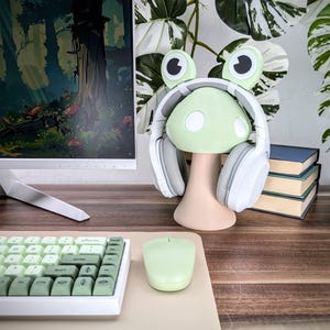 May include: A green frog-shaped headphone stand with white earcups and a mushroom-shaped base. The stand is on a wooden surface with a keyboard and a mouse.