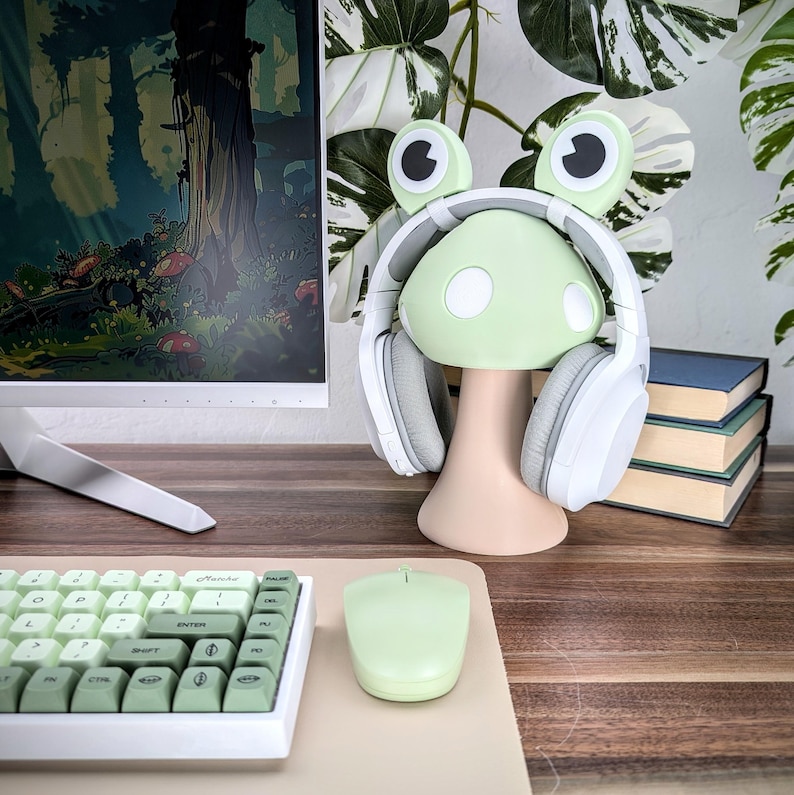 May include: A green frog-shaped headphone stand with white earcups and a mushroom-shaped base. The stand is on a wooden surface with a keyboard and a mouse.