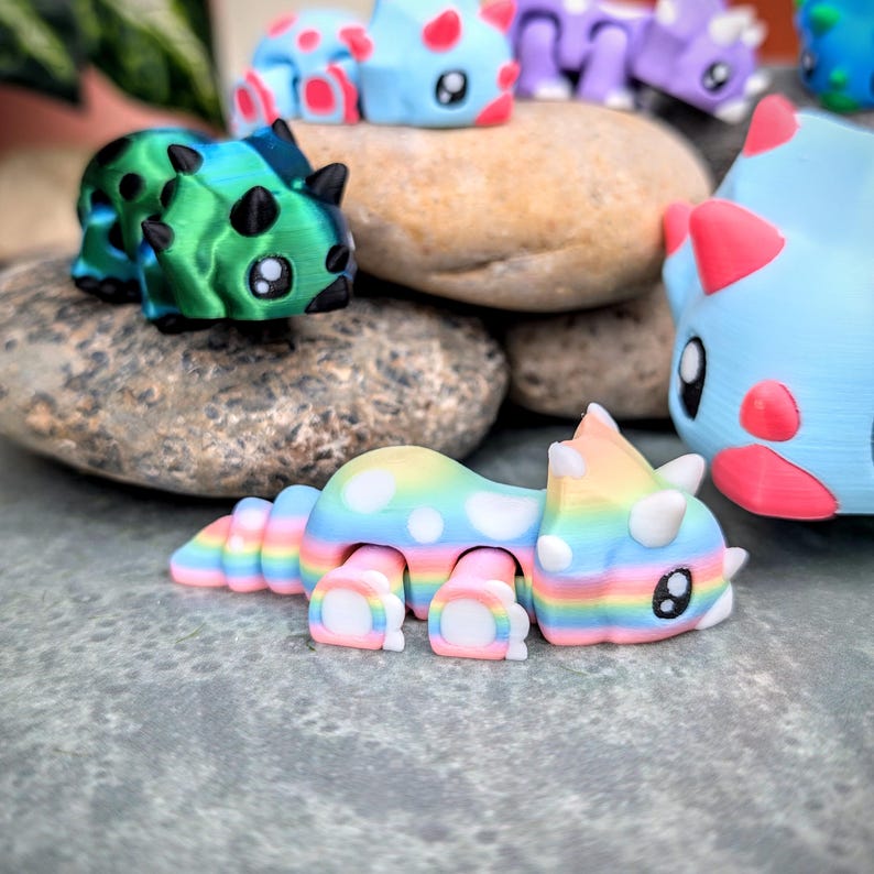 May include: A rainbow-colored, 3D printed dinosaur toy with white polka dots. The dinosaur is lying on its belly with its head turned to the side. The toy is made of a soft, flexible material.