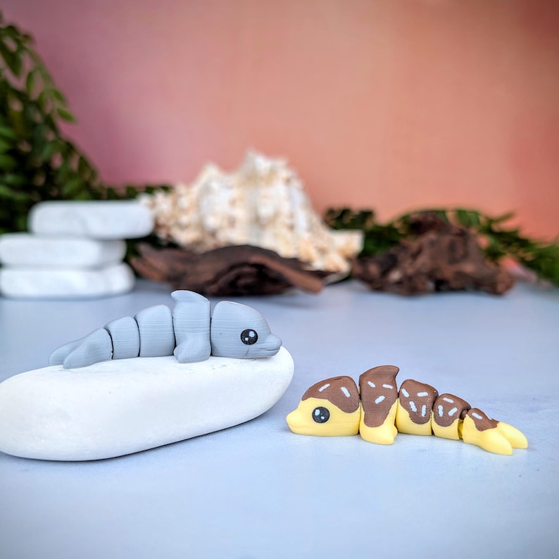 May include: Two articulated dolphin figurines. One is gray and resting on a white stone. The other is yellow and brown. The background includes a seashell, wood, and greenery, with a pink and blue backdrop.