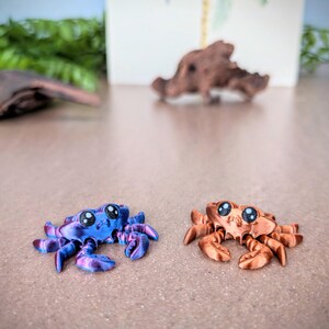 May include: Two small, 3D printed crabs. One is a metallic blue and purple color, the other is a metallic copper color. Both have large, black eyes.