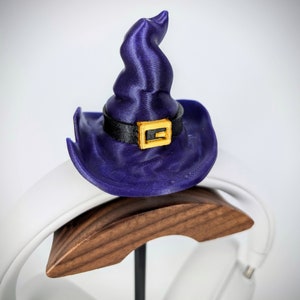 May include: A purple witch hat with a gold buckle sits on a wooden headphone stand. The stand is white and has a curved shape.