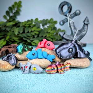 May include: A selection of brightly coloured shark figurines, including pink, blue, and rainbow, displayed on small stones. A decorative anchor is in the background. The sharks have cartoonish features and are arranged on a light blue surface.