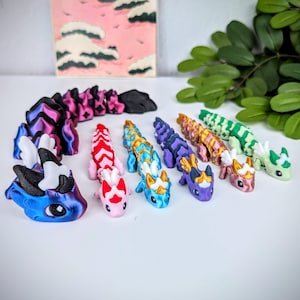 K&ouml;nnte beinhalten: Eine Sammlung farbenfroher, beweglicher Drachenfiguren in verschiedenen Farben, darunter Blau, Rosa, Rot, Gold, Lila und Gr&uuml;n. Jeder Drache hat detaillierte Merkmale und ein einzigartiges Design. Der Hintergrund umfasst ein Gem&auml;lde und Gr&uuml;n.