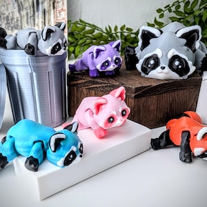 May include: A set of five small, colorful, articulated raccoon figurines. The raccoons are posed in different positions, including sitting, lying down, and standing. The figurines are made of a smooth, plastic material.