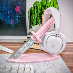 May include: A pink headset stand shaped like a katana sword, holding a white headset. The stand has a pink base and a silver blade. The headset is white with gold accents.