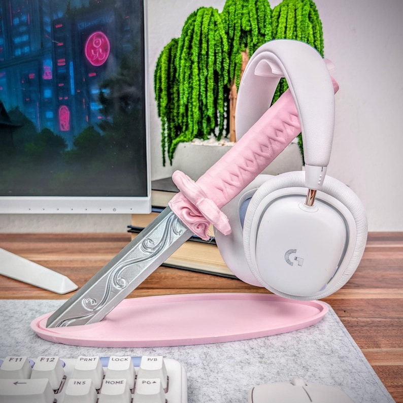 May include: A pink headset stand shaped like a katana sword, holding a white headset. The stand has a pink base and a silver blade. The headset is white with gold accents.