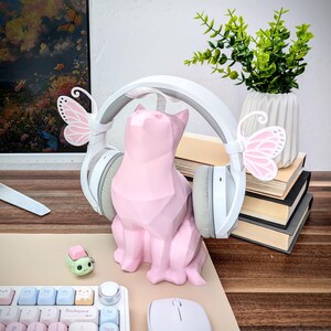 May include: A pink geometric cat figurine with white headphones and pink butterfly accents on each earcup. The cat is sitting on a wooden surface with a keyboard and mouse in the background.