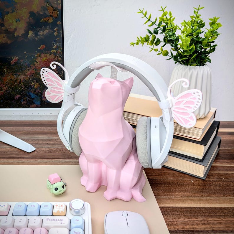 May include: A pink geometric cat figurine with white headphones and pink butterfly accents on each earcup. The cat is sitting on a wooden surface with a keyboard and mouse in the background.