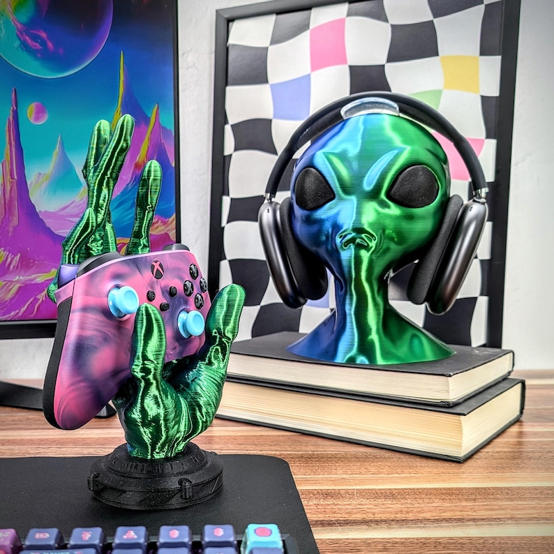 May include: A green alien head with headphones, a pink and black video game controller, and a green hand holding the controller. The alien head is on a stack of books. The controller is on a black stand.