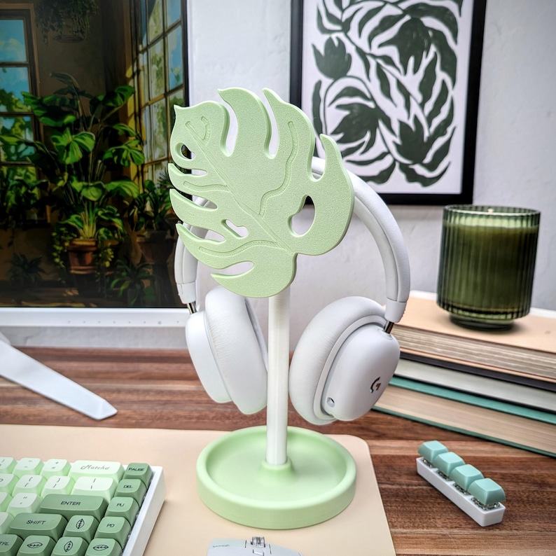 May include: A light green monstera leaf shaped headphone stand with a white headphone resting on it. The stand is on a light brown wooden surface with a keyboard and a mouse in the background.
